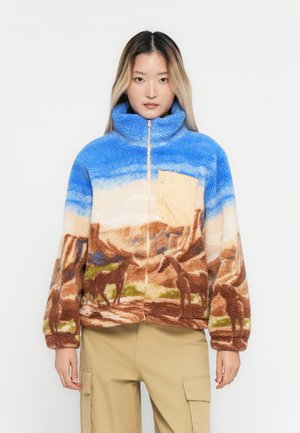 Kvinne iført en blå og brun fleecejakke med et ørkenlandskap og hestemønster, beige brystlomme og beige cargo-bukser.