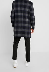 Abrigo de cuadros en lana azul marino y gris con una silueta larga y una tela ligeramente texturizada; acompañado de pantalones ajustados negros y zapatillas blancas.