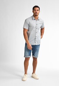 Camicia a maniche corte con bottoni di colore grigio chiaro con motivi blu; abbinata a pantaloni shorts in denim arrotolati e sneakers color crema.