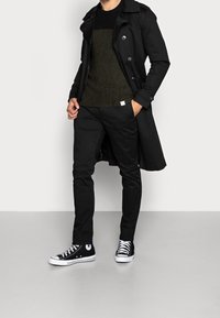 Svart trenchcoat över en mörkgrön stickad tröja, kombinerad med svarta byxor och svartvita sneakers. Har knappar och texturkontrast.