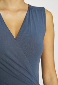 Top senza maniche blu con scollo a V, caratterizzato da un design avvolgente e un tessuto liscio. Nessun motivo visibile o dettagli in metallo.