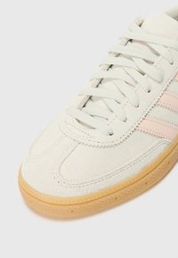 Biały zamszowy trampek z jasnoróżowymi paskami, płaskimi sznurowadłami i gumową podeszwą. Charakteryzuje się gładką teksturą i przeszyciami.