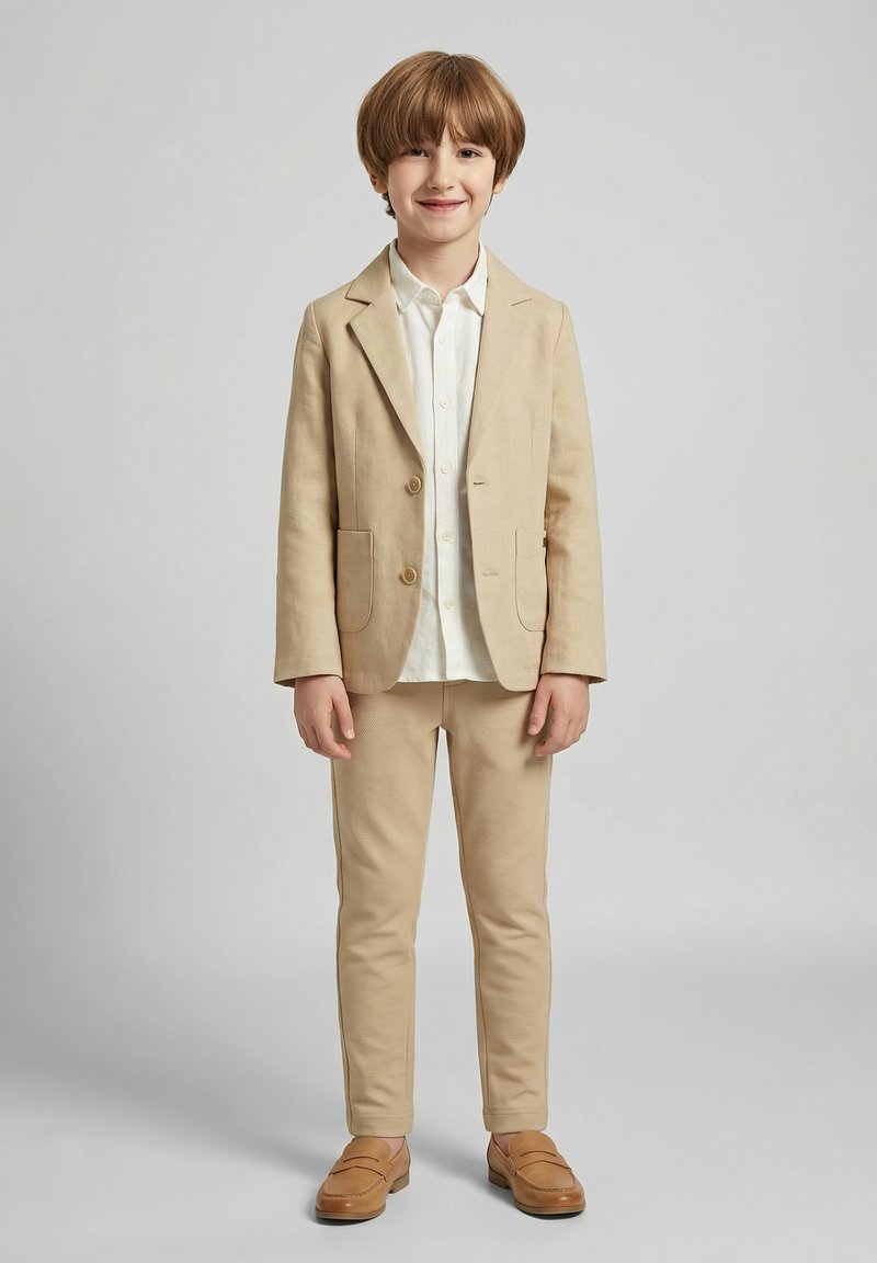 Jonge jongen die glimlacht, gekleed in een beige colbert, bijpassende broek, wit overhemd met knopen en beige loafers, staand tegen een effen lichte achtergrond.