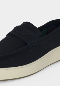 Chaussure en daim noir à enfiler avec bout mocassin et semelle en caoutchouc blanche, avec détail de bride cousue sur le dessus.