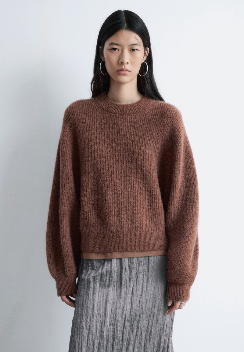 Pull texturé marron avec un col rond et des épaules tombantes, associé à une jupe plissée grise. Design minimaliste, matériau chaud.