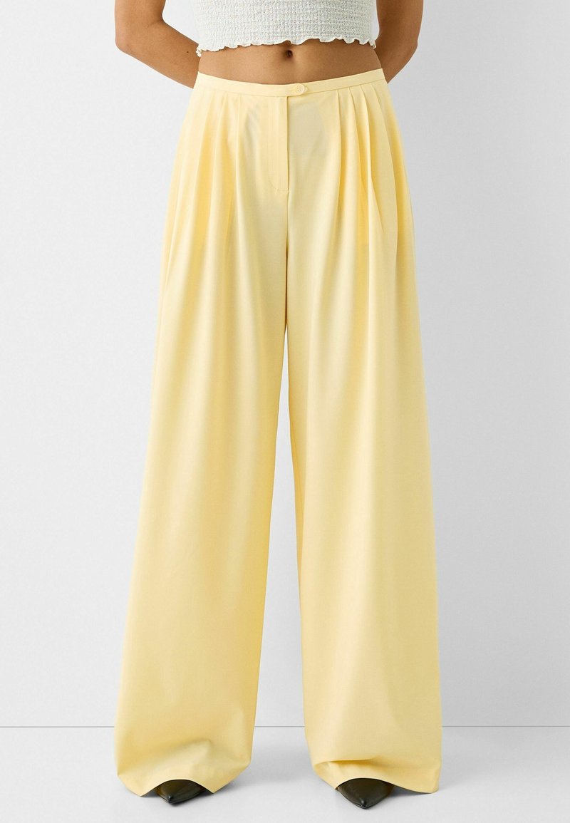 Wide-leg trousers in light yellow fabric, featuring front pleats and a single button closure. Smooth texture and a relaxed fit.