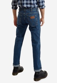 Jeans de mezclilla azul con un corte recto, dobladillos enrollados y un parche de cuero en la parte posterior. Cuenta con dos bolsillos traseros con costuras decorativas.