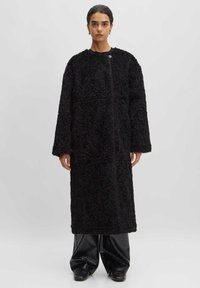 Zwarte, harige jas met een lang, oversized ontwerp, met een ronde halslijn en een sluiting aan de voorkant, in combinatie met glanzende zwarte wijd-pijp broeken.