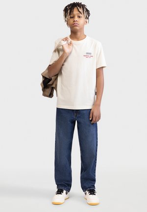 Αγόρι με ανοιχτό καφέ μπουφάν περασμένο στον ώμο, φορώντας μπεζ μπλούζα, μπλε τζιν και λευκά αθλητικά παπούτσια, στέκεται μπροστά σε λευκό φόντο.