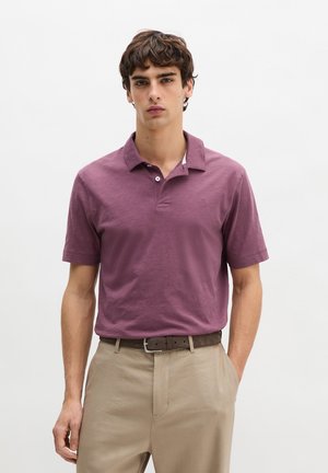 Junger Mann, der ein kurzärmeliges Poloshirt in Mauve trägt, in beige Hosen mit einem braunen Gürtel, stehend vor einem schlichten weißen Hintergrund.