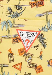 Tela amarilla con motivos de palmeras y gráficos de temática de viajes. Incluye un parche de logo triangular rojo de GUESS® en el bolsillo.