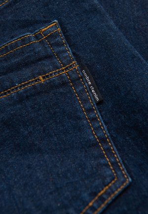 Tessuto di denim scuro con cuciture arancioni, con una piccola etichetta nera con la scritta "ombre denim" vicino al bordo di una tasca, che mostra una texture liscia.