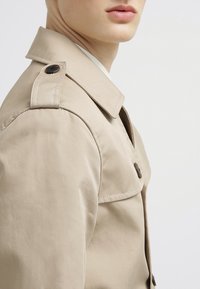 Gros plan d'une personne portant un trench beige avec une bride d'épaule boutonnée et un col, sur un fond clair uni.
