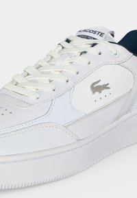 Λευκό αθλητικό παπούτσι Lacoste με λευκά κορδόνια, ασημί λογότυπο κροκόδειλου στο πλάι και σκούρο μπλε εσωτερική επένδυση ορατή στη φτέρνα.
