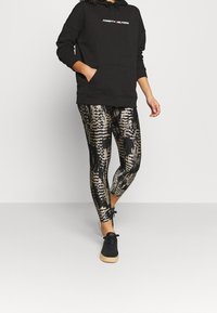 Felpa nera con tasca frontale e logo grafico; leggings a fantasia con tonalità oro e nere; abbinata a sneakers nere.