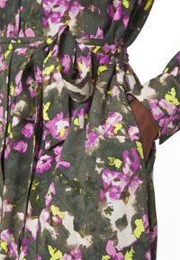 Robe à motif floral avec des fleurs violettes, jaunes et blanches sur tissu vert foncé, dotée d'une ceinture nouée à la taille et d'une main dans la poche latérale.