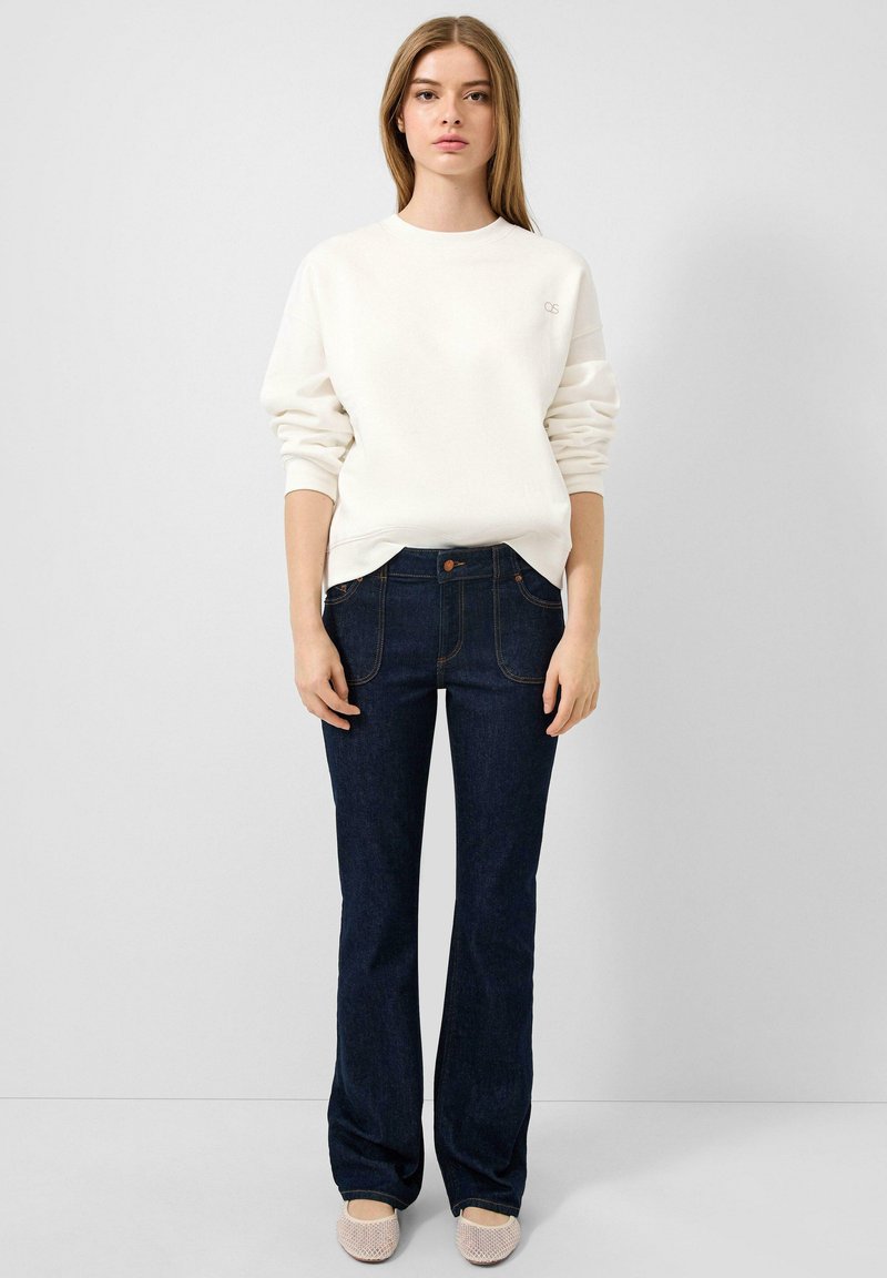 Witte crewneck sweatshirt, donkerblauwe flare jeans met voorkruiken en lichte instappers. Zachte textuur, casual design.