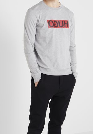 Mann trägt einen grauen Langarmpullover mit rotem und schwarzem "HUGO"-Schriftzug und schwarze Hose, die Hände in den Taschen, steht vor weißem Hintergrund.