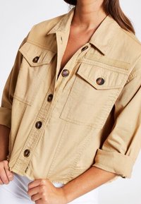 Tan välsydd jacka i bomullstyg, med två bröstfickor, fem runda knappar och uppvikta ärmar. Slät textur.