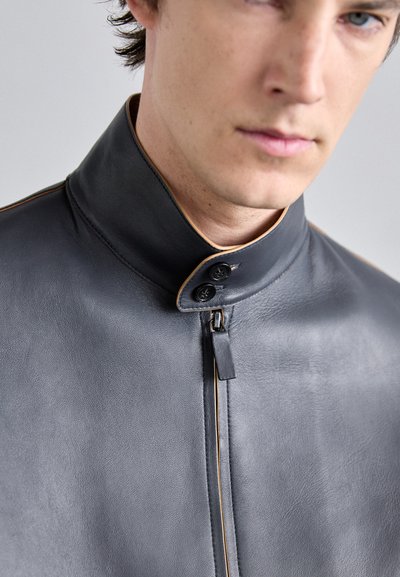 Schwarze Lederjacke mit hohem Kragen, ausgestattet mit einem Reißverschluss vorne, zwei Knöpfen und kontrastierenden Paspeln entlang des Kragens und des Reißverschlusses.
