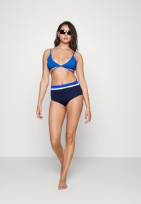 Top de bikini azul com design entrelaçado, combinado com parte de baixo alta em azul-marinho com risca de acento branca e azul. Modelo a usar óculos de sol.