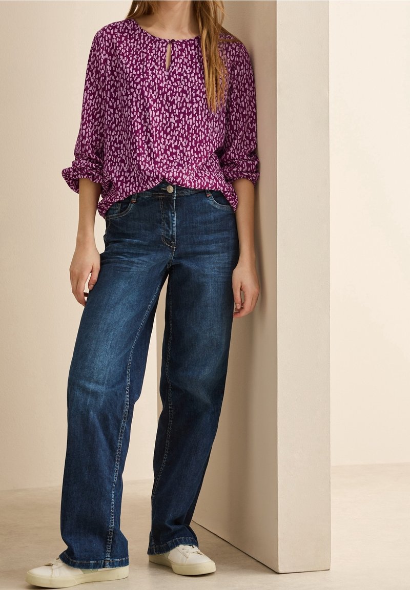 Femme portant un chemisier violet à motifs, un jean droit bleu foncé et des baskets blanches, debout et appuyée contre un mur beige.