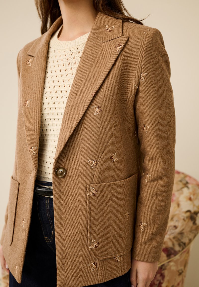 Bruine houndstooth blazer met vlinderborduursel, enkele knoopsluiting en twee steekzakken aan de voorkant. Draagt over een crèmekleurige gebreide trui.