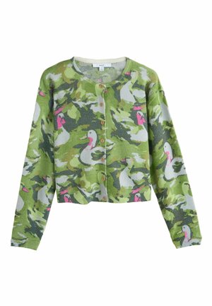 Giovane donna in piedi con le mani nelle tasche, indossa un cardigan mimetico verde, pantaloni beige a gamba larga e sneakers dorate su sfondo grigio.
