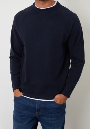 Homme portant un pull à manches longues bleu marine sur une chemise blanche, les mains dans les poches d'un jean bleu foncé, debout devant un fond uni.