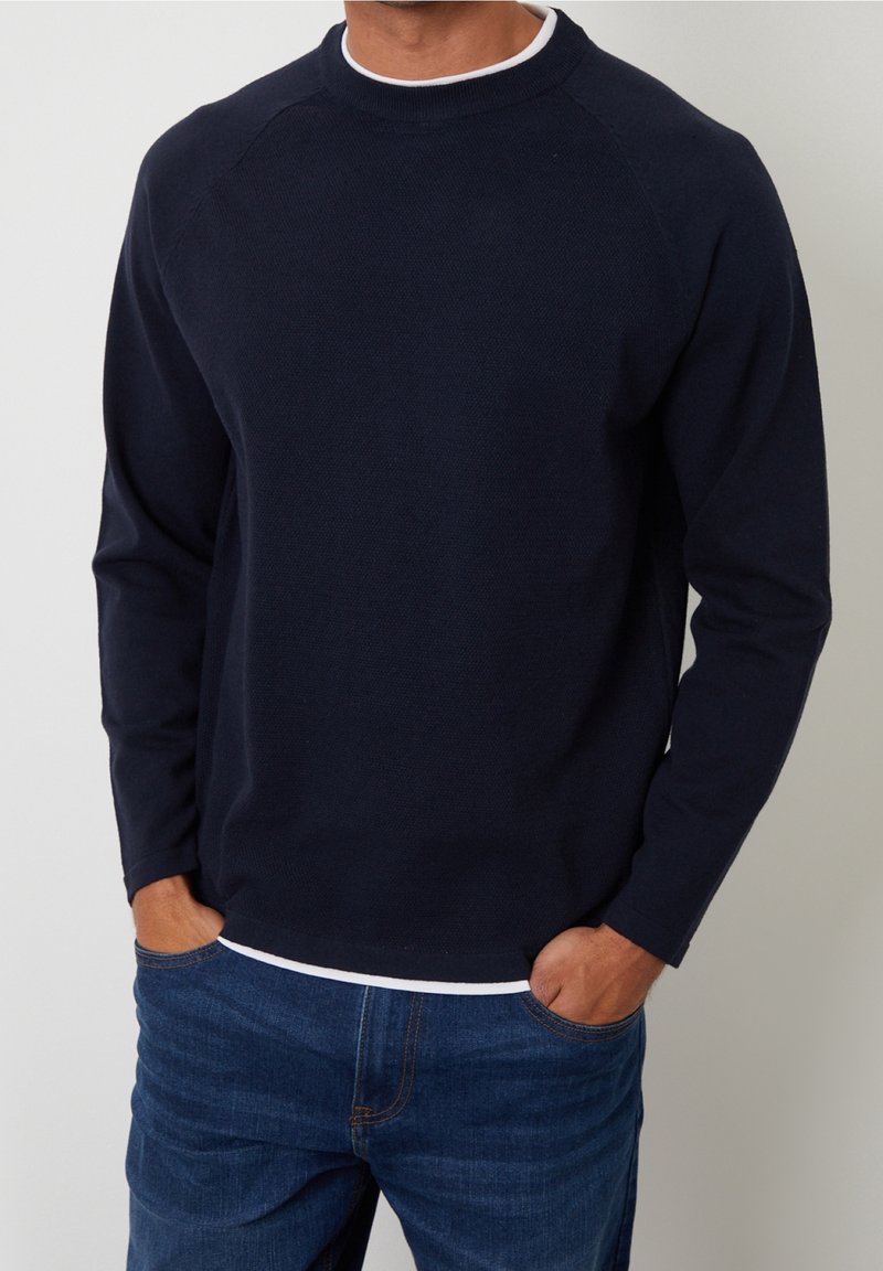 Homme portant un pull à manches longues bleu marine sur une chemise blanche, les mains dans les poches d'un jean bleu foncé, debout devant un fond uni.