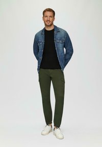 Jeansjack met twee borstzakken, zwart shirt met ronde hals, olijfgroene cargobroek en witte sneakers, staand tegen een neutrale achtergrond.