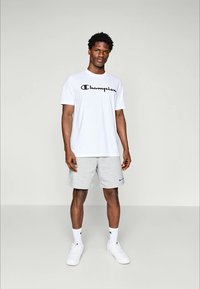 Weißes T-Shirt mit kurzen Ärmeln und schwarzem "Champion"-Logo sowie grauen Sportshorts. Die Accessoires umfassen weiße Socken und Sneaker.