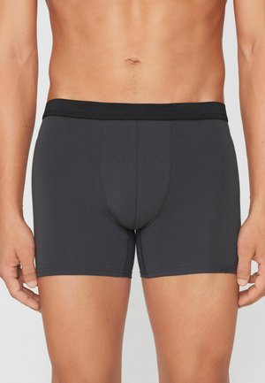 Mannelijke torso met effen donkergrijze boxershort met zwarte tailleband, staand tegen een neutrale achtergrond.
