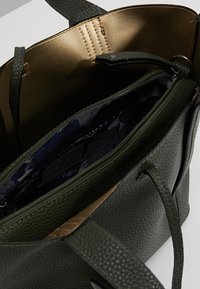 Borsa tote in verde oliva con texture e interno dorato, dotata di tasca con zip e fodera in tessuto con un motivo a foglia. Include spallacci regolabili.