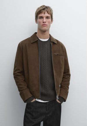 Jonge man die een bruine suède jas draagt over een donkere kabelgebreide trui en zwarte broek, staande tegen een effen witte muur.