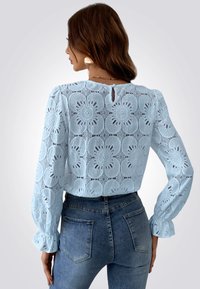 Top de encaje azul claro con estampado floral, mangas largas y puños con volantes; la parte posterior presenta un pequeño ojal con cierre de botón.