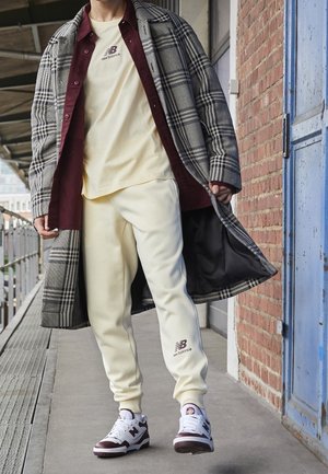 Osoba ubrana w kremowy dres New Balance, białe i bordowe buty sportowe, bordową koszulę oraz szary płaszcz w kratę stoi przy ścianie z cegieł obok niebieskich drzwi.