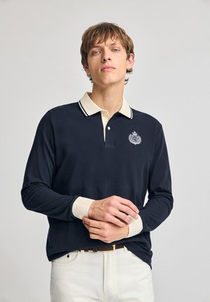 Navy long-sleeve polo shirt with cream collar and cuffs; features embroidered logo and contrasting striped details on the collar.