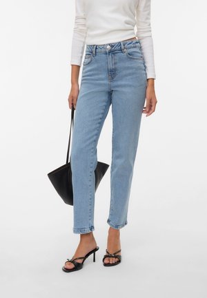 Femme portant un jean taille haute bleu clair, un haut blanc à manches longues, des sandales noires à talons et tenant un sac à main noir à côté d'elle.