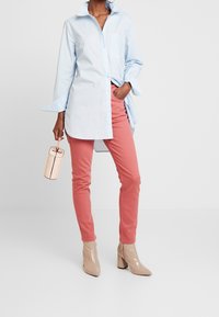 Camisa azul claro de gran tamaño con cuello puntiagudo, combinada con pantalones ceñidos en color coral. La modelo sostiene un bolso redondo de color durazno y lleva botas de tacón alto beige.