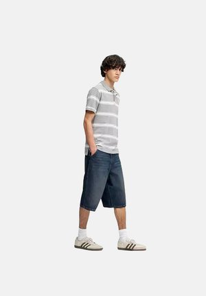 Ung mand går med hænderne i lommerne iført en gråstribet polo, mørke lange shorts, hvide sokker og beige sneakers.