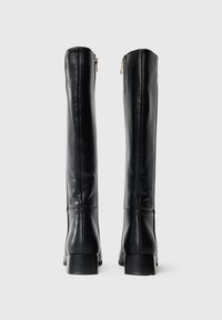 Bottes en cuir noir montant jusqu'aux genoux, avec une finition lisse, une forme structurée et un petit talon bloc. Dotées d'une fermeture éclair à l'arrière pour un enfilage facile.