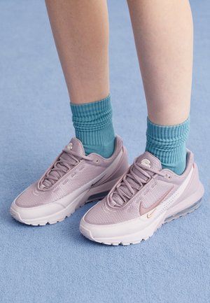 Baskets de sport lilas clair avec une tige en mesh et un amorti Air visible, accompagnées de chaussettes côtelées teal. Posées sur un fond textured bleu.