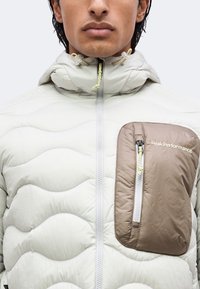 Homme portant une doudoune de couleur claire avec col montant et poche poitrine étiquetée PeakPerformance avec fermeture éclair.