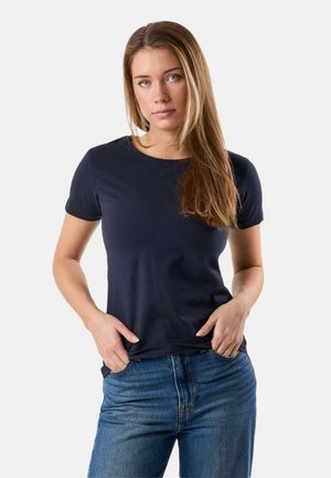 Jeune femme aux longs cheveux châtain clair portant un t-shirt bleu marine et un jean bleu, debout les mains dans les poches devant un fond blanc.