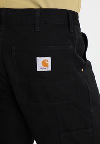 Zwarte werkbroek met een achterzak met een witte vierkante patch en oranje logo. Gemaakt van duurzaam materiaal met zichtbare stikseldetails.