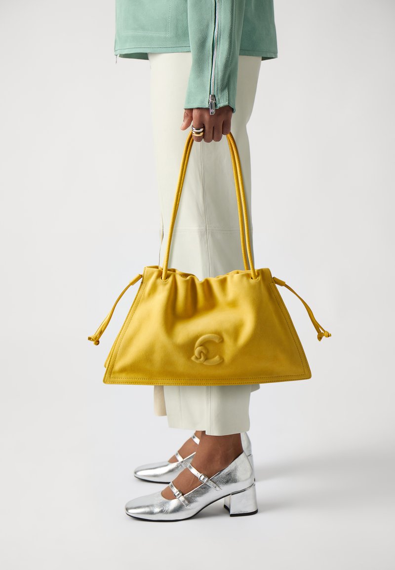 Sac à main en daim jaune avec un cordon de serrage, arborant un logo texturé. Porté avec un pantalon crème et des chaussures à talons carrés argentés.
