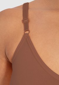 Spallina di reggiseno in tessuto marrone con fibbia regolabile e dettaglio ad anello. Taglio a triangolo e texture liscia, vista ravvicinata sulla pelle.