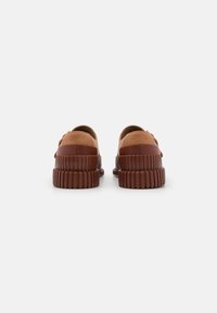 Brązowe gumowe buty z grubymi, żłobionymi podeszwami, z gładką górą i wytłoczonym logo z tyłu, ogólnie minimalistyczne detale.