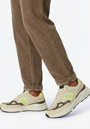 Persoon die beige sneakers met neonaccenten en dikke witte zolen draagt, samen met bruine corduroybroek, staand op een witte achtergrond.